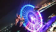 People are enjoying a nighttime swing ride against the backdrop of a brightly lit Ferris wheel and city lights.