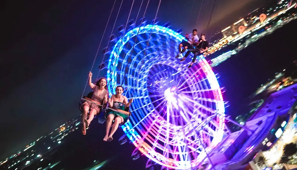 People are enjoying a nighttime swing ride against the backdrop of a brightly lit Ferris wheel and city lights