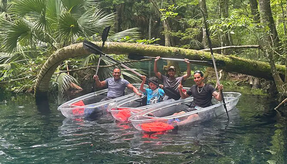 Four people are smiling and posing with their paddles in clear kayaks on a calm transparent waterway amidst lush greenery