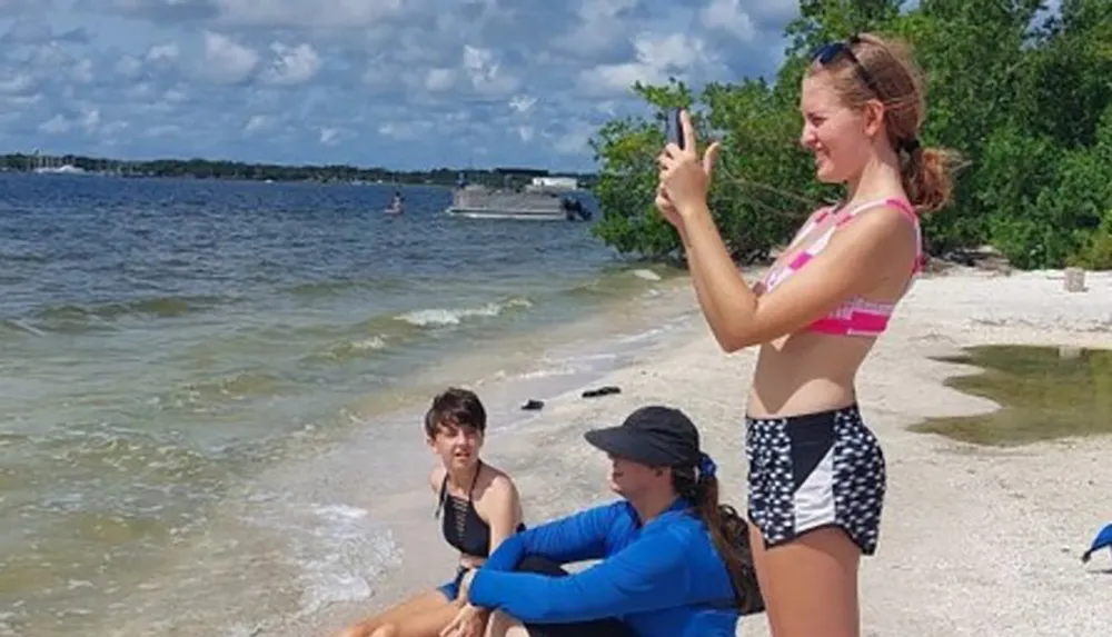 A person is standing and taking a selfie on the beach while two others sit by the waters edge