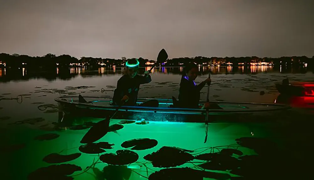 Two people are kayaking at night on a body of water illuminated with a bright green light from beneath revealing underwater vegetation