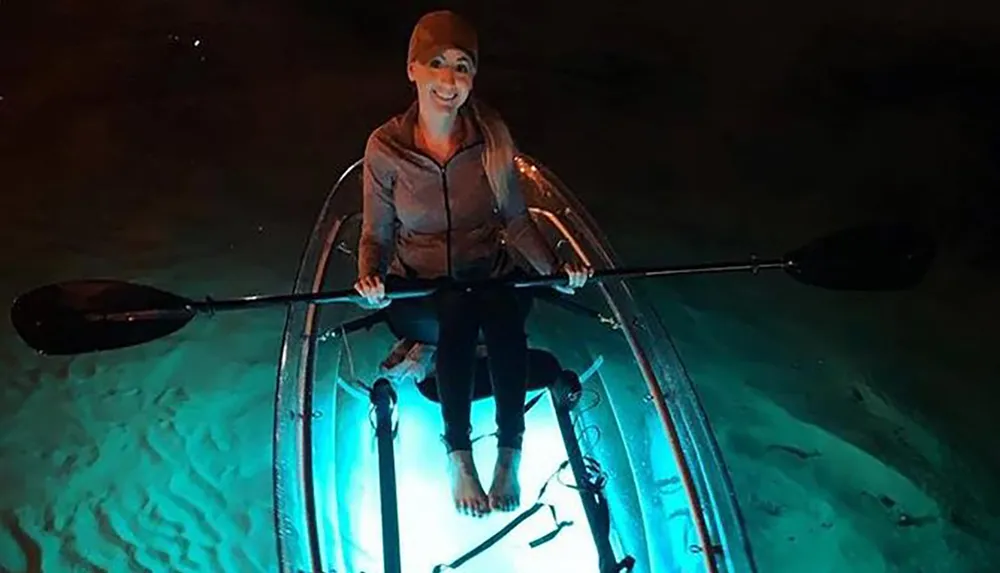 A person is kayaking at night in a clear kayak illuminated from within highlighting the water below