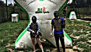 Two people wearing masks and protective vests are holding paintball guns, standing next to an inflatable barrier in a paintball arena.