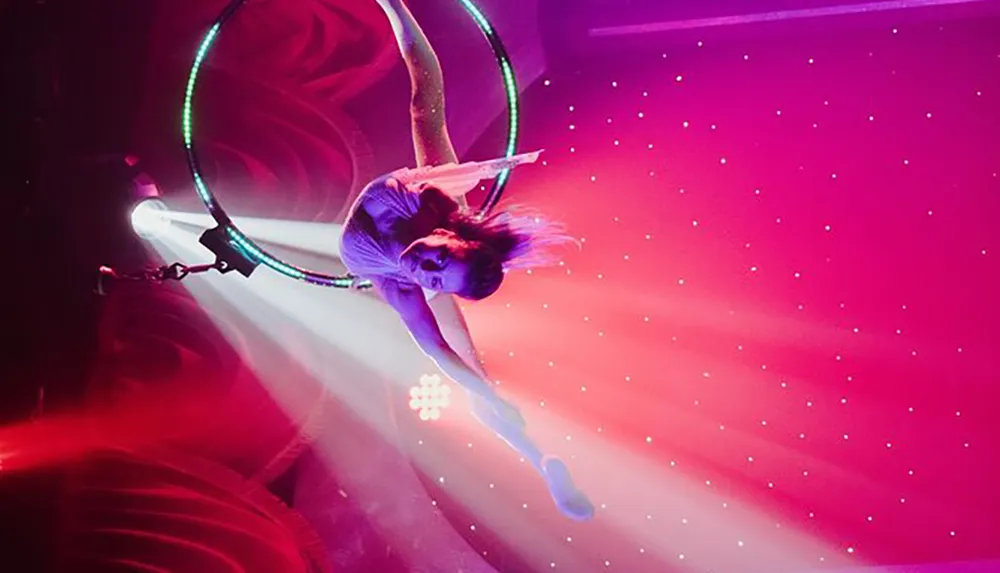 An aerial performer is suspended in mid-air executing an acrobatic pose on a hoop against a vibrant backdrop of pink and purple lights