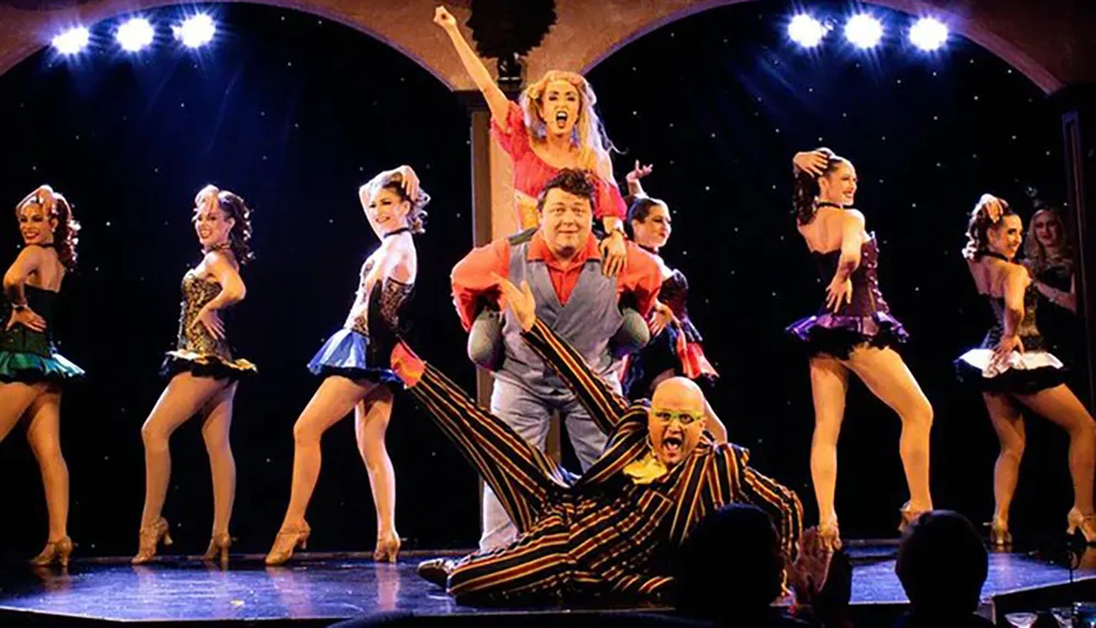 A vibrant theatrical performance featuring dancers in colorful costumes and two central characters engaging in comedic antics on stage