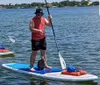 A person wearing a life jacket is paddleboarding on calm water with a rabbit aboard the board