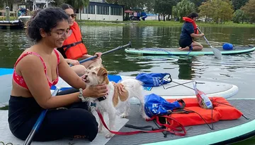 A person is smiling as they sit on a paddleboard with a happy-looking dog, while others paddle nearby on what appears to be a calm body of water.