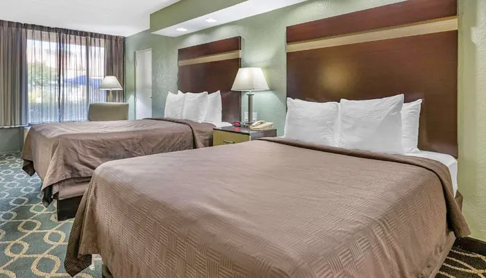 The image shows a neatly arranged hotel room with two double beds nightstands with lamps and a window with curtains