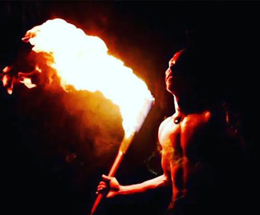A person is performing a fire-breathing act against a dark background producing a large flame from their mouth