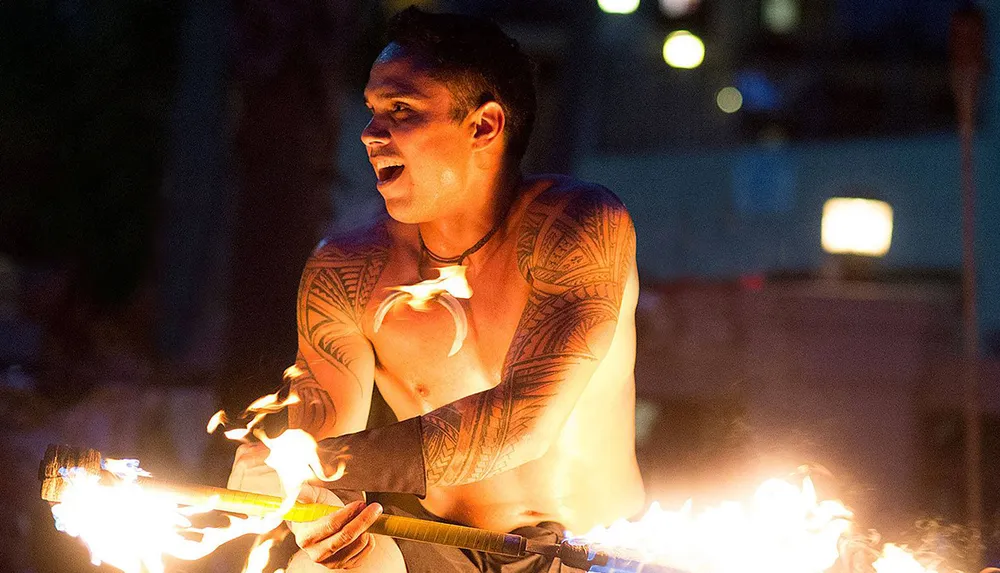 A performer with traditional tattoos smiles as he adeptly manipulates a flaming staff at a nighttime event