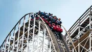 Thrilled riders are descending a steep drop on a wooden roller coaster against a clear sky.