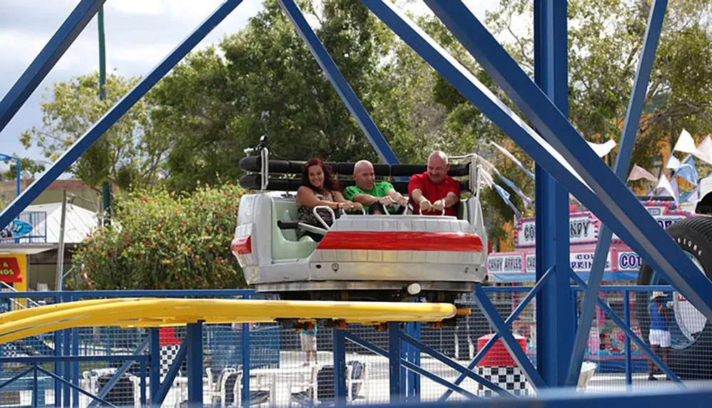 Three people enjoy a ride on a small roller coaster in an amusement park setting