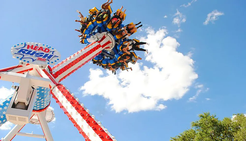 Thrill-seekers are strapped into a spinning amusement park ride called the Head Rush 360 flying through the air against a backdrop of blue sky and fluffy clouds