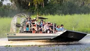 A group of tourists wearing headphones is enjoying a high-speed ride on an airboat through a marshy landscape.