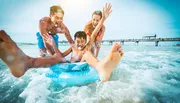 A group of joyful people is having fun in the sea with a young person on an inflatable ring, taking in the sun near a pier.