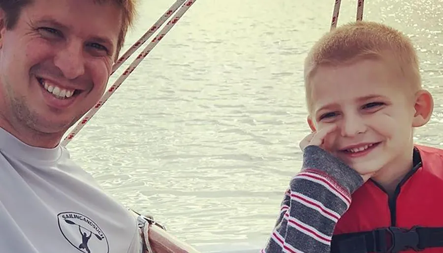 A man and a young boy are smiling at the camera with a backdrop of water, possibly out on a boat.