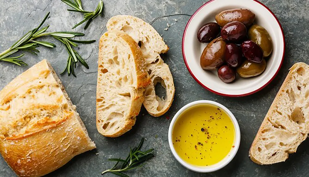 The image shows a simple and appetizing arrangement of sliced bread a bowl of mixed olives olive oil with pepper and fresh rosemary on a textured surface