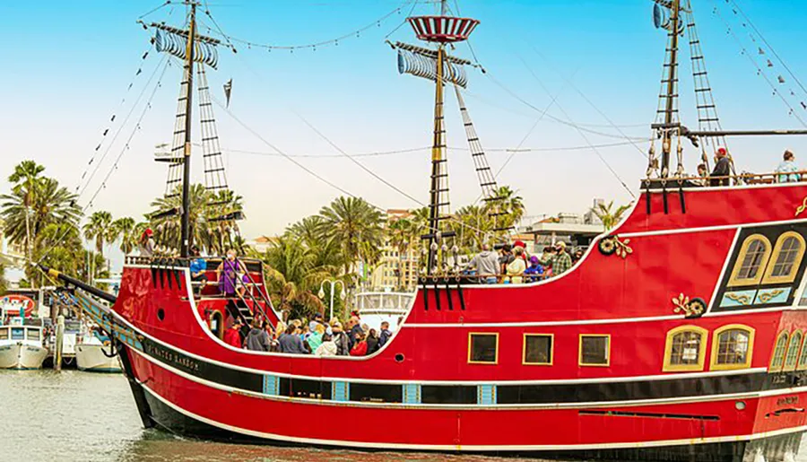 A red pirate-themed tour boat is docked at a harbor, bustling with passengers on board and visitors around in a tropical setting with palm trees in the background.