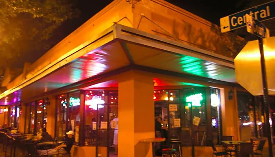 An illuminated street corner cafe at night showcases colorful lights under its awning and patrons enjoying the outdoor seating.