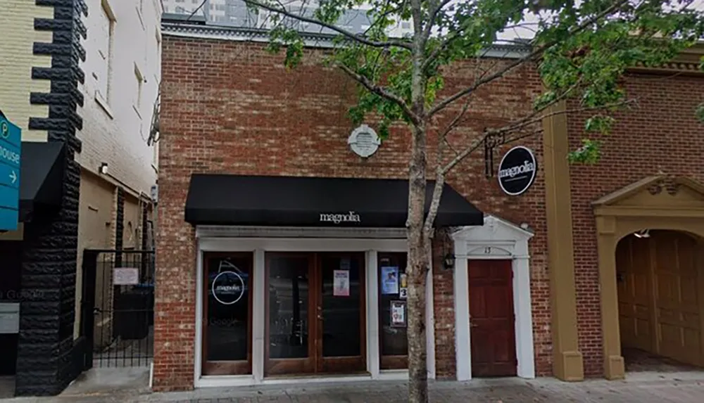 The image shows a storefront with a black awning labeled magnolia a sidewalk tree in front and adjacent buildings on a calm street