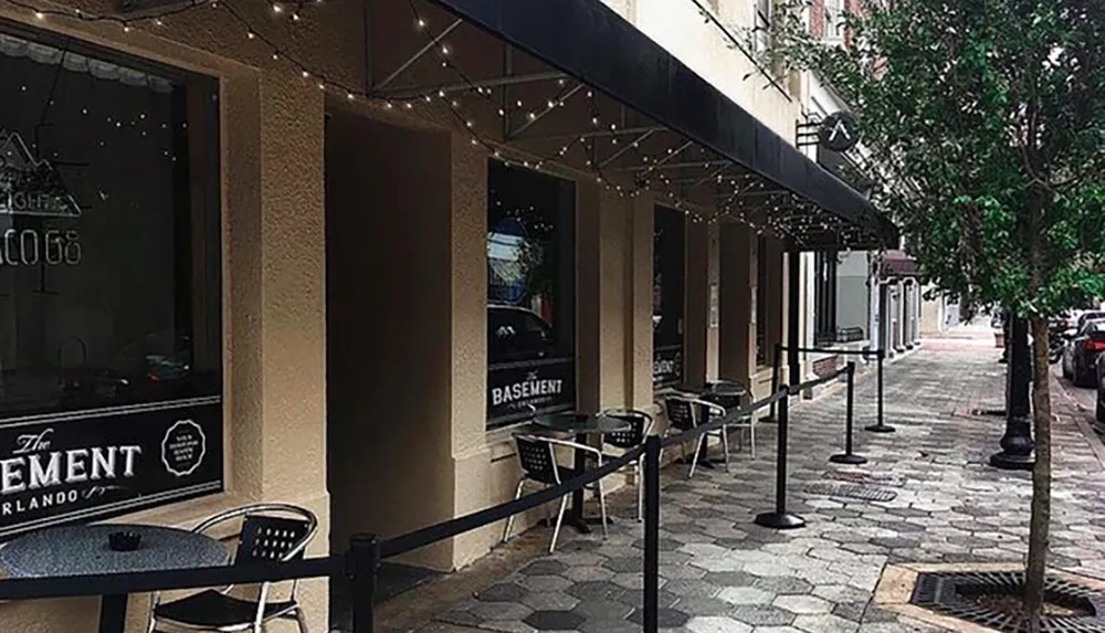 A quiet sidewalk cafe setting with string lights under an awning in an urban environment