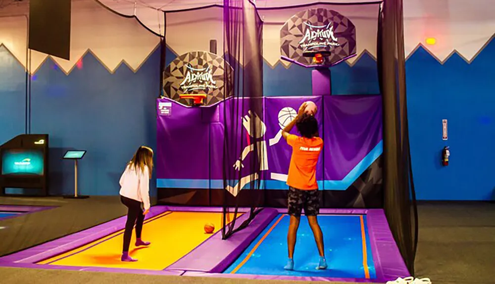 A person in an orange shirt attempts a basketball shot on an indoor court with trampoline flooring while another person watches