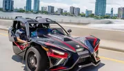 A person is driving a three-wheeled Polaris Slingshot on a bridge with a city skyline in the background.
