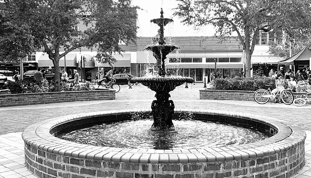 This black and white image captures a three-tiered fountain surrounded by a brick circular border situated in a public space with trees people bicycles and the backdrop of a commercial area