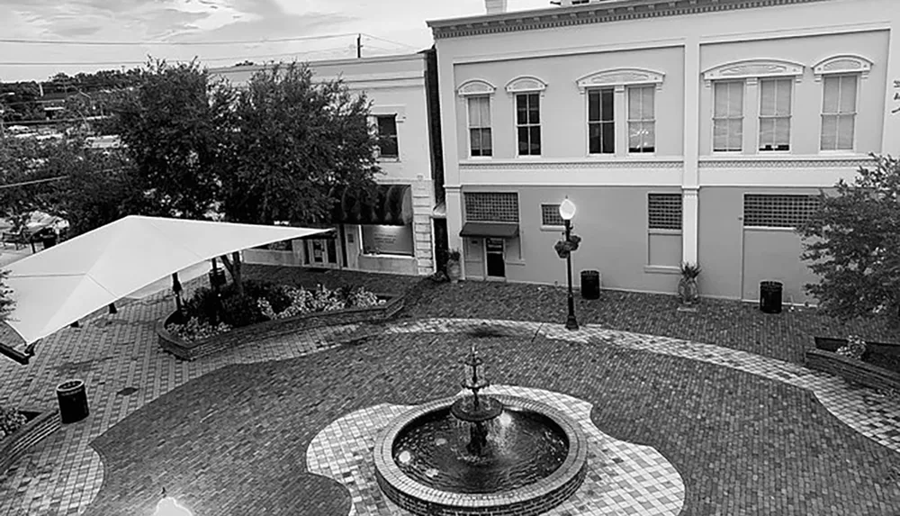 The black and white photo depicts a quaint urban plaza with a circular brick pattern a fountain in the center an awning to the left buildings in the background and no visible people