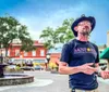 A man wearing a cowboy hat and a Sanford Tours  Experiences t-shirt is speaking while gesturing with his hands standing in an outdoor plaza with a fountain and string lights in the blurred background