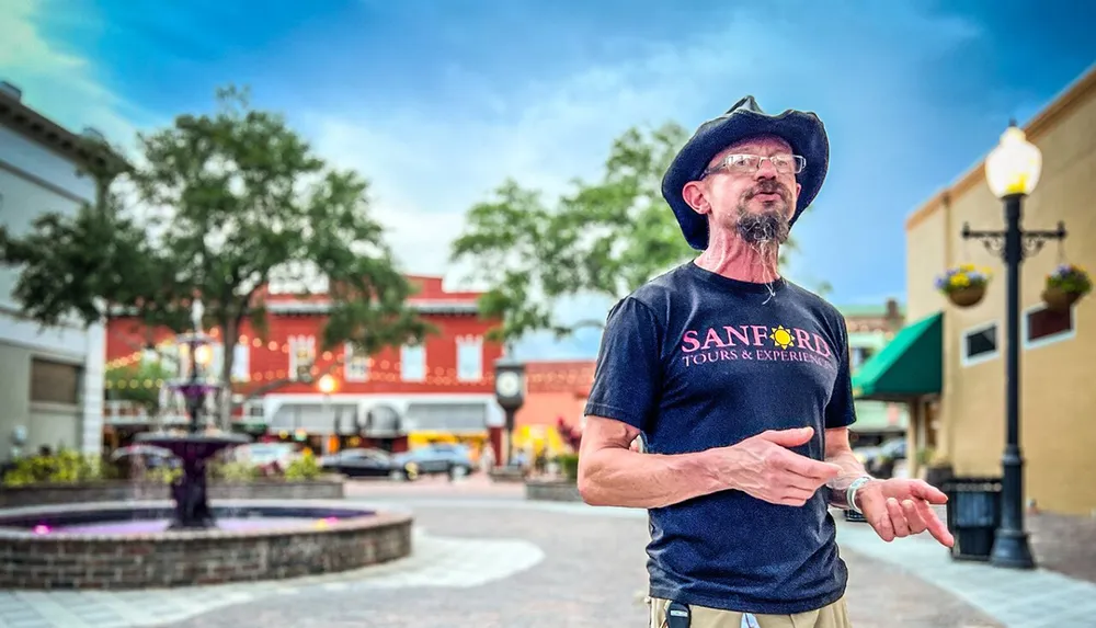 A man wearing a cowboy hat and a Sanford Tours  Experiences t-shirt is speaking while gesturing with his hands standing in an outdoor plaza with a fountain and string lights in the blurred background
