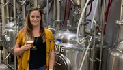 A woman is posing with a glass of beer in front of stainless steel brewing tanks in a brewery.