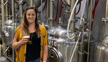 A woman is posing with a glass of beer in front of stainless steel brewing tanks in a brewery.