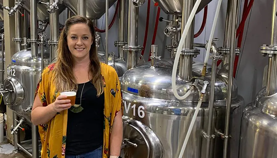 A woman is posing with a glass of beer in front of stainless steel brewing tanks in a brewery.