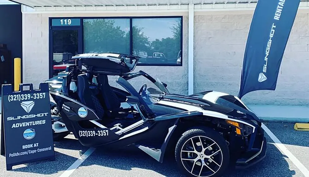 A black Polaris Slingshot three-wheeled vehicle is on display outside a rental establishment with advertising banners and a sign promoting adventures and bookings