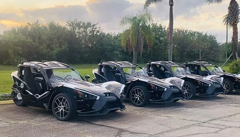 The image shows a lineup of Polaris Slingshot three-wheeled vehicles parked side by side with a backdrop of palm trees and a cloudy sky