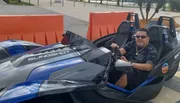 A person is seated in a parked Polaris Slingshot three-wheeled vehicle, smiling and holding the steering wheel.