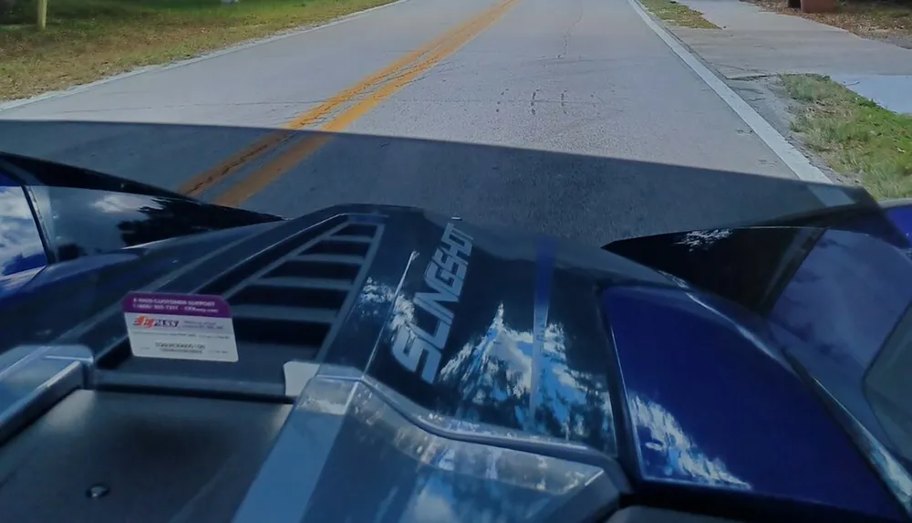 The image shows the perspective of a driver in a Polaris Slingshot vehicle indicated by the Slingshot writing on the hood gazing down a tree-lined road with a clear sky reflected on the vehicles surfaces