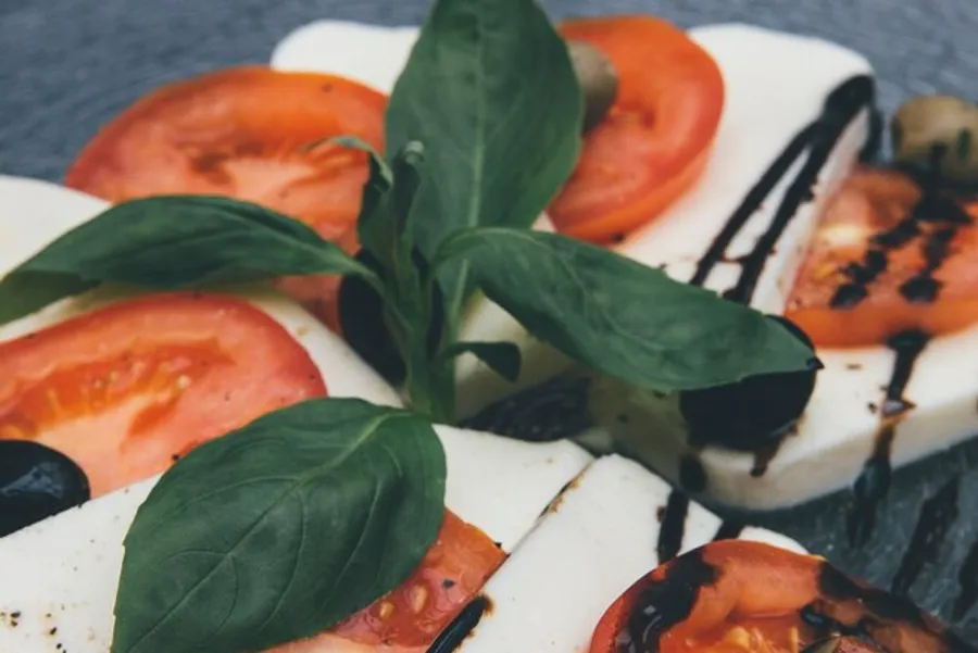 The image shows a caprese salad with alternating slices of tomato and mozzarella cheese, garnished with fresh basil leaves and drizzled with balsamic reduction.