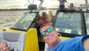 A group of people is enjoying a boat ride on a sunny day, with some sitting at the back and one person taking a selfie in the foreground.