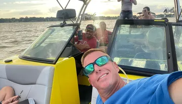 A group of people is enjoying a boat ride on a sunny day, with some sitting at the back and one person taking a selfie in the foreground.