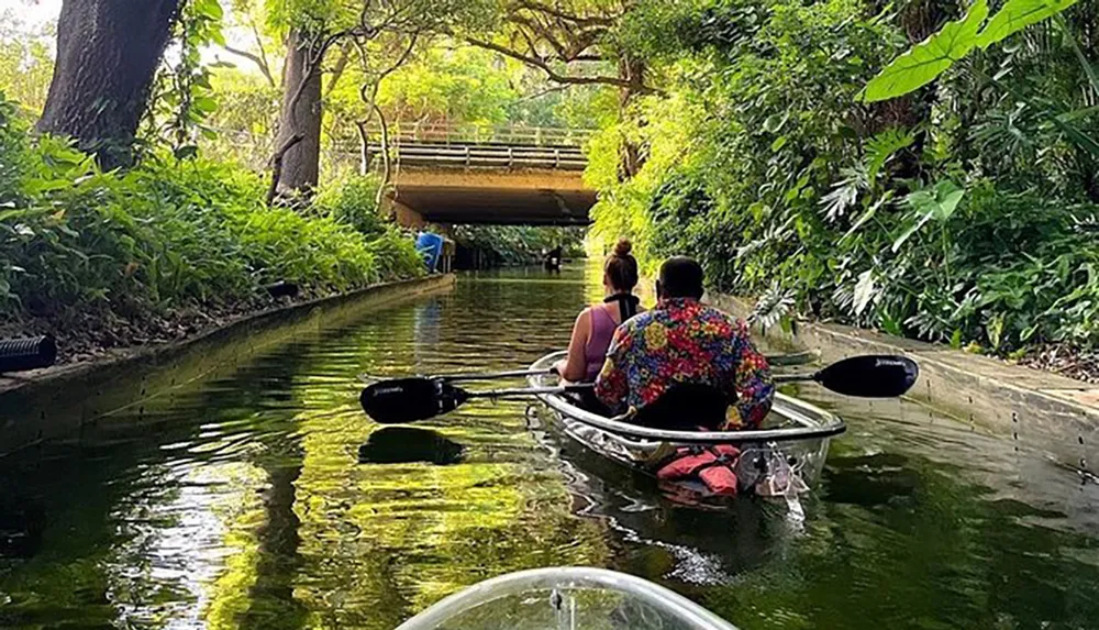 Two people are kayaking down a peaceful tree-lined canal with a bridge in the background