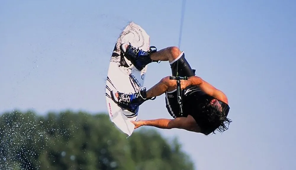 A wakeboarder is captured in mid-air performing a trick with the wakeboard flipped upwards and their body in an inverted position against a clear blue sky