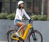 A person is seated on a yellow electric bicycle with a rear cargo rack holding the handlebars and appears ready to ride