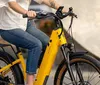 A person is seated on a yellow electric bicycle with a rear cargo rack holding the handlebars and appears ready to ride