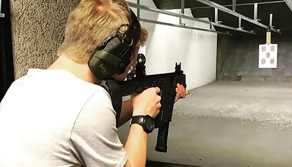 A person wearing protective headphones is aiming a rifle at a target in an indoor shooting range