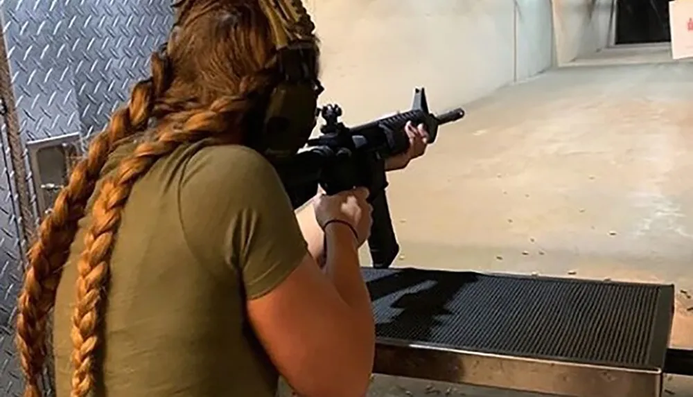 A person with a long braid is aiming a rifle at a target at an indoor shooting range