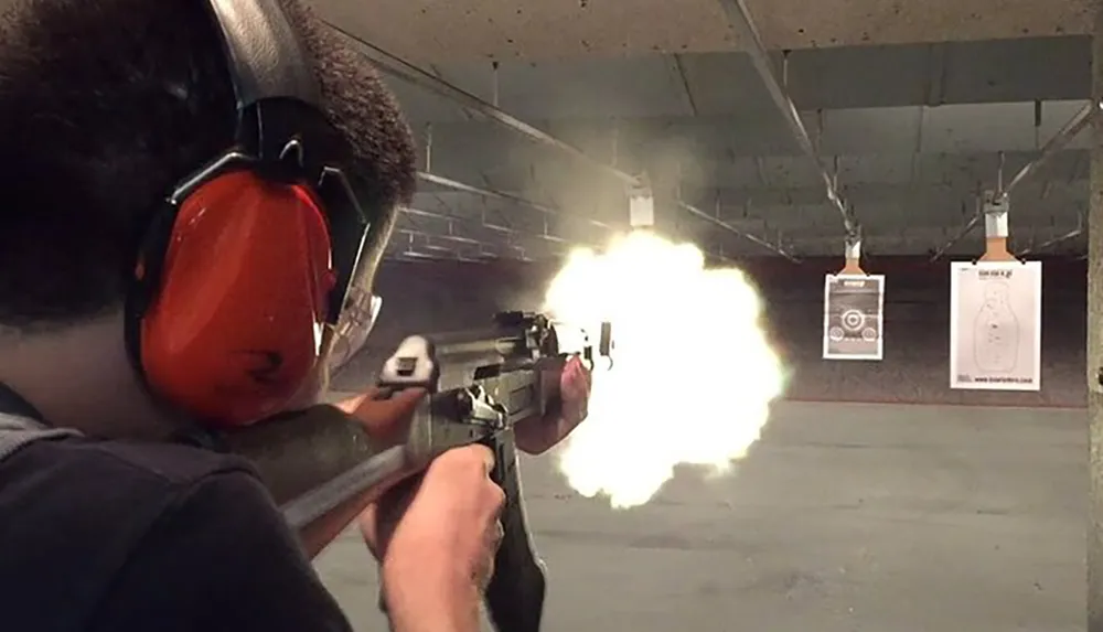 A person is firing a rifle at a shooting range with a visible muzzle flash and target papers in the background
