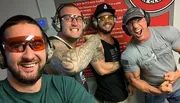 Four men are smiling and posing for a selfie, wearing protective eyewear and headphones typically used for ear protection, suggesting they are at a shooting range.