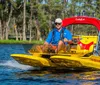 A man wearing a life jacket and sunglasses is steering a yellow compact boat with the label CraigCat through a body of water with greenery and trees in the background